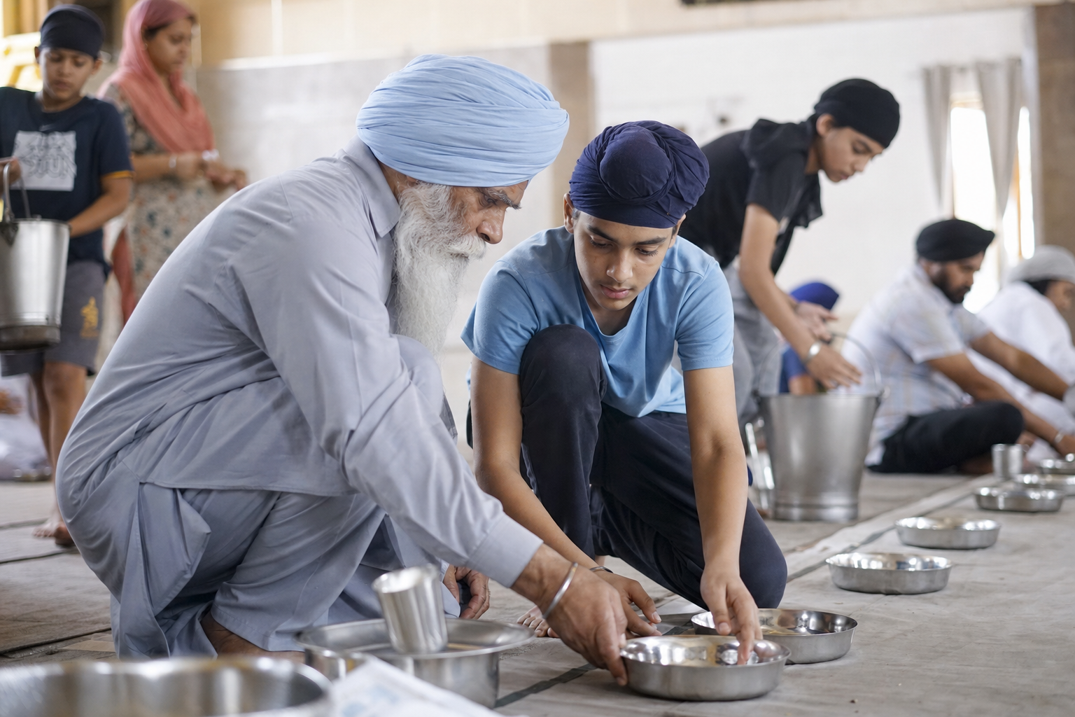 An elderly Sikh volunteer shows a teenager how to arrange steel bowls while people of different ages prepare a langar hall.