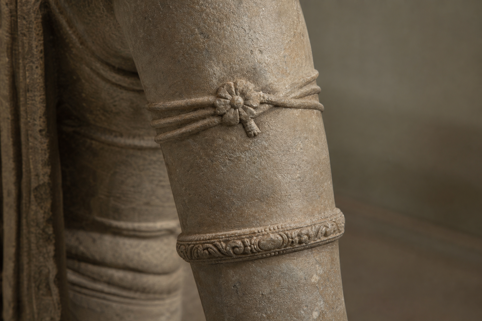 Close-up of a carved upper arm with three tied cords, a floral clasp, and a separate rigid bracelet below.