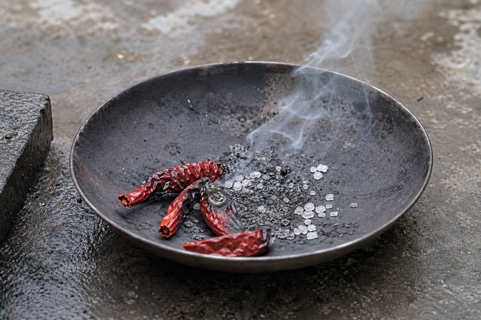 A faint irregular curl of smoke disperses above dried chillies and salt in a shallow metal vessel outdoors.