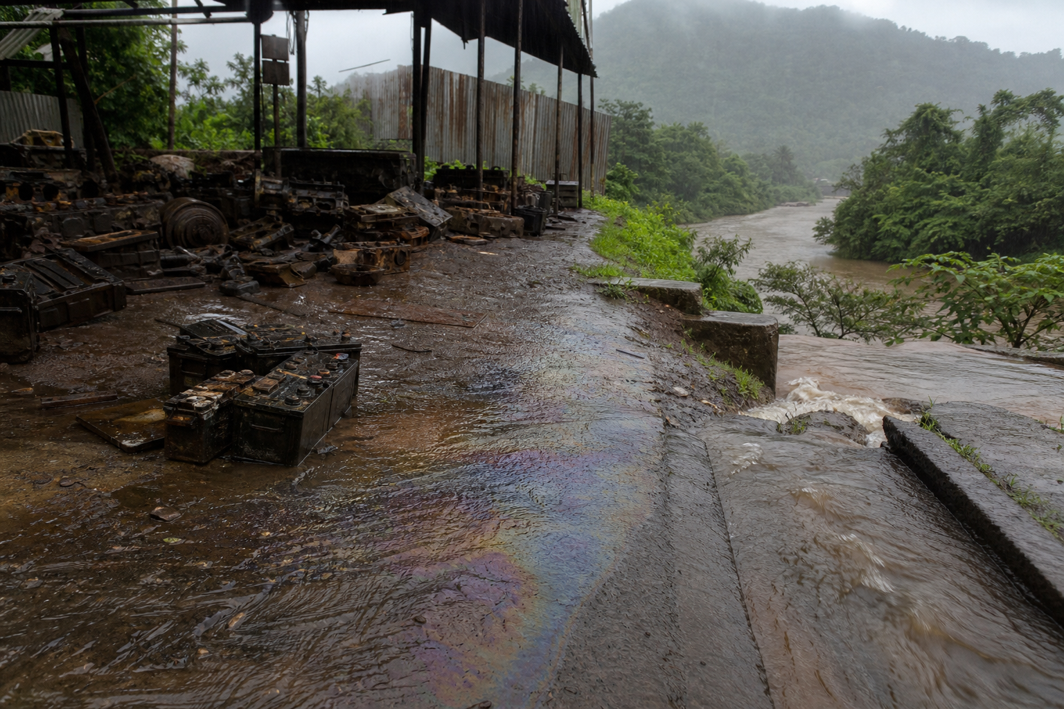 Monsoon runoff flows from uncovered scrap and machinery parts into a roadside drain near a green watercourse in Chiplun.