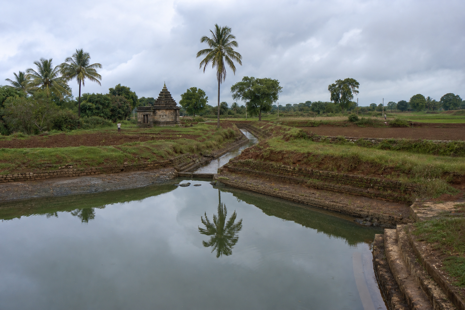 A village tank and earthen watercourse lead through cultivated fields toward a weathered stone temple at Devarabelakere.
