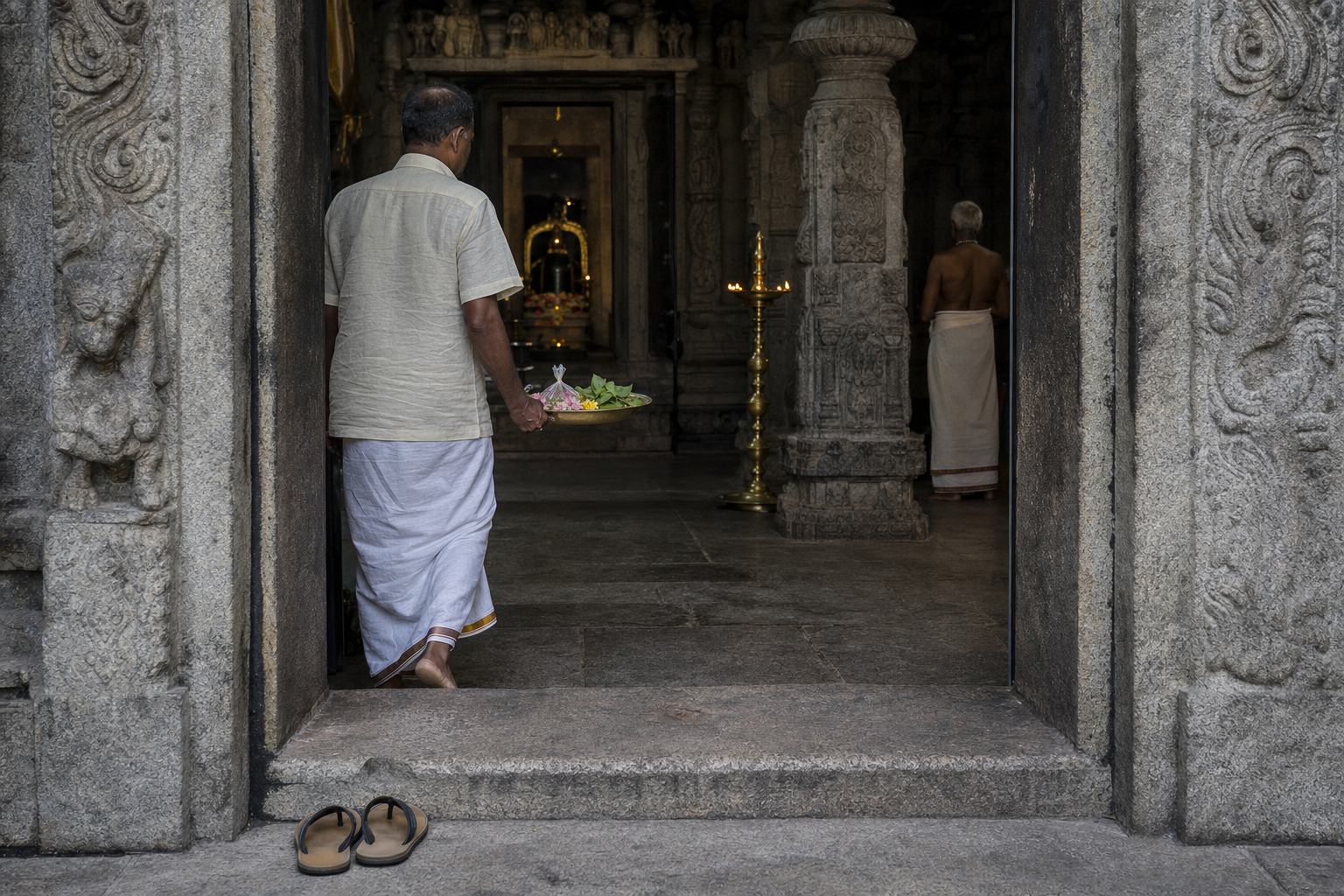 A devotee carrying flowers and bilva leaves enters a shaded stone temple passage while another worshipper waits near a lamp.