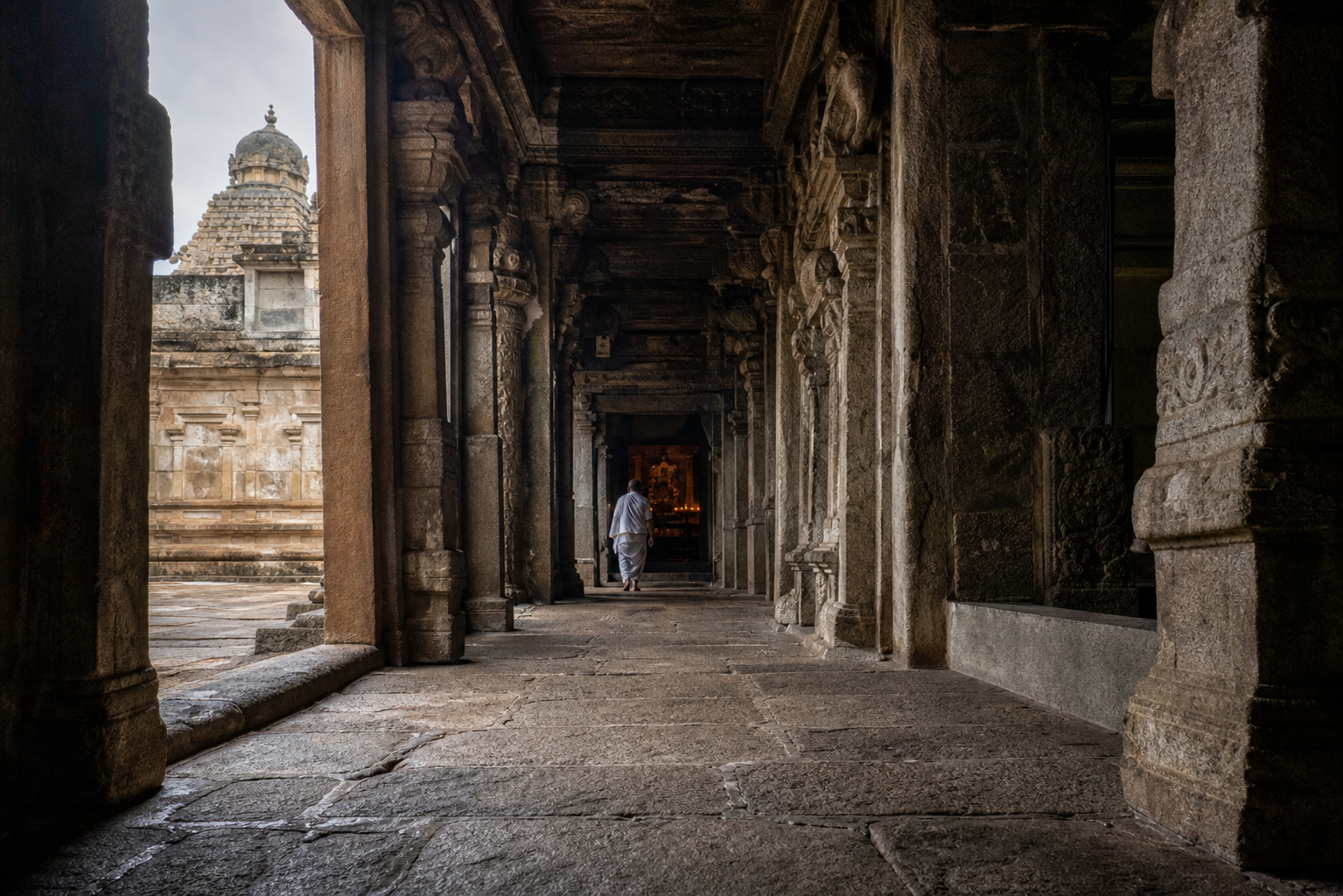 A worn granite path passes through pillared temple spaces toward a lamp-lit sanctum doorway, with a devotee walking ahead.
