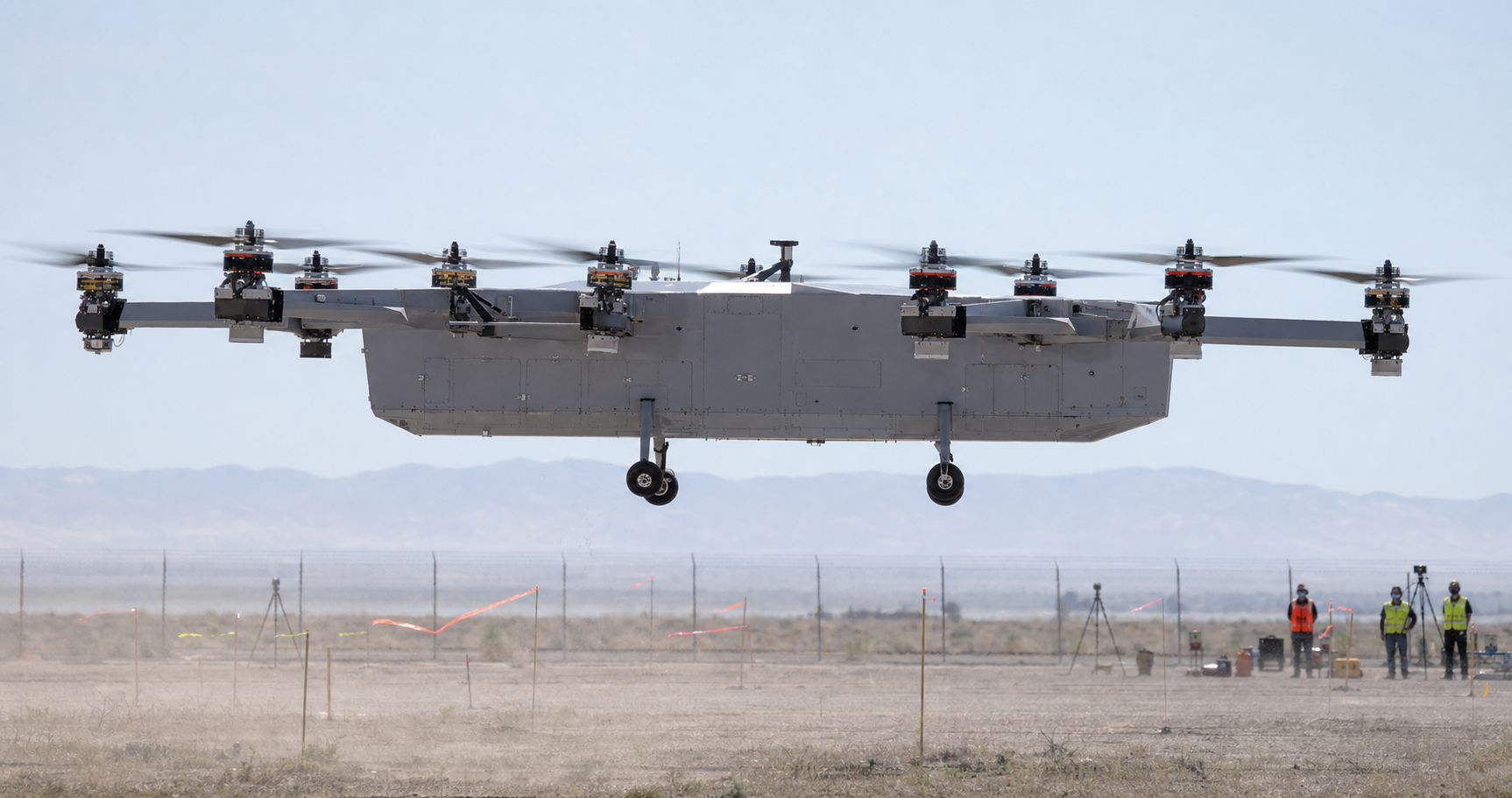 A large instrumented electric VTOL demonstrator hovers level inside a secured test range as cameras and a remote crew monitor the flight.