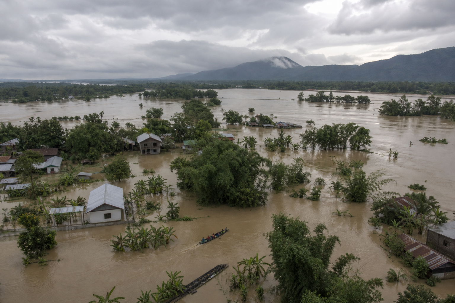 Floodwater surrounds homes, fields and trees on Assam's Brahmaputra floodplain as a small rescue boat travels toward higher ground beneath monsoon clouds.