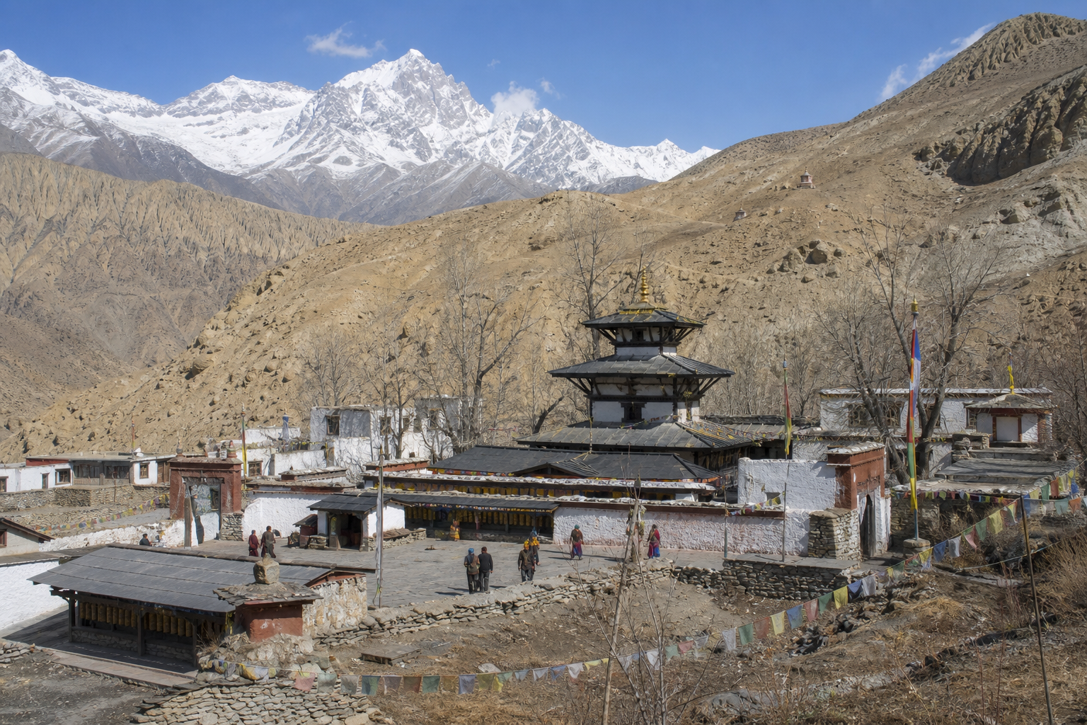 The pagoda-roofed temple and whitewashed religious buildings at Muktinath stand beneath dry Himalayan slopes and distant snowy peaks as pilgrims cross the courtyard.
