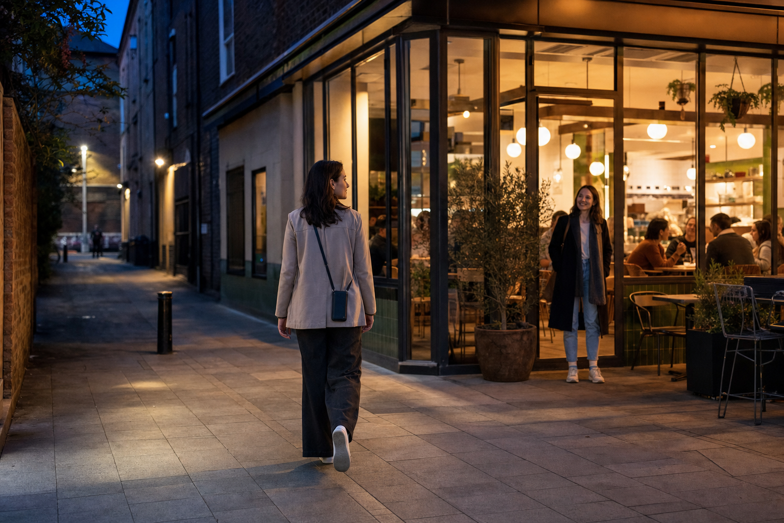 A woman chooses a busy, well-lit pavement and walks toward a waiting companion during an evening journey.