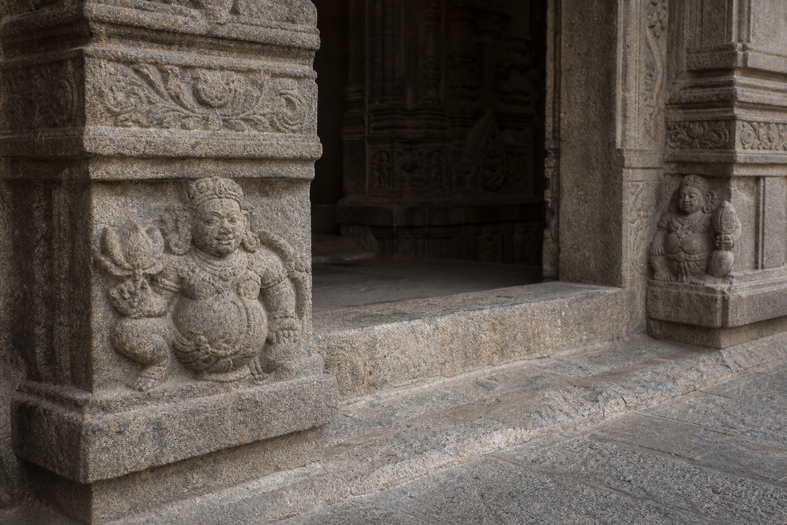 Lower temple doorway showing a lotus-bearing pot-bellied figure opposite a conch-bearing companion within the carved threshold ensemble.