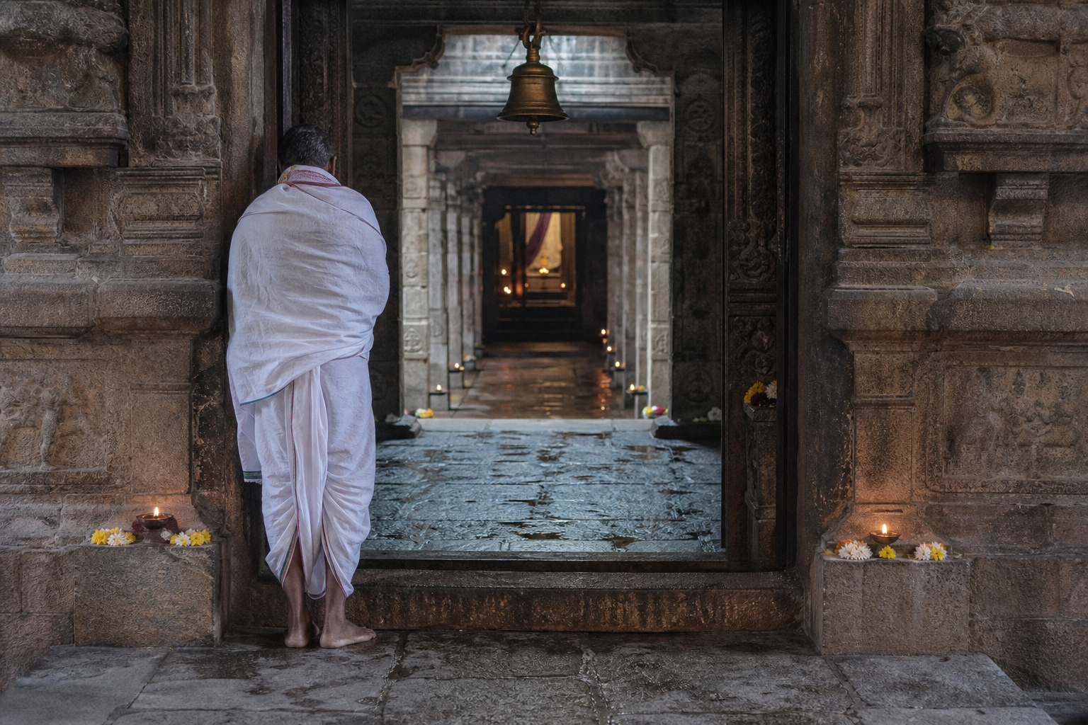 A barefoot pilgrim pauses at a carved temple threshold leading through pillared halls toward a softly lit sanctum.