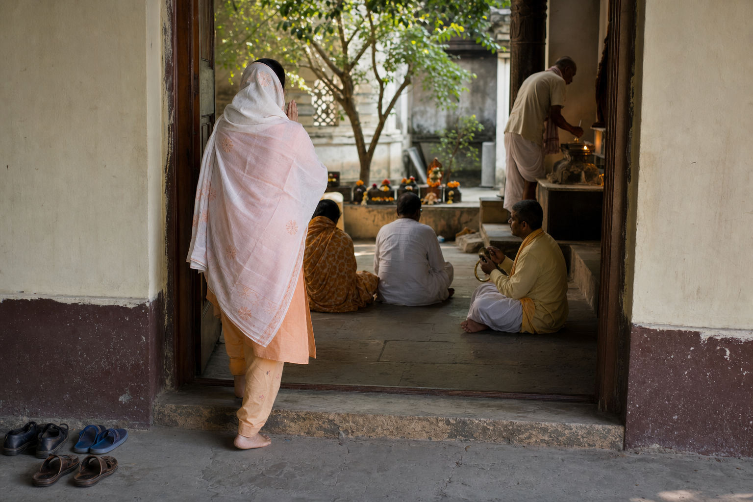 A barefoot pilgrim with folded hands enters a shaded sacred courtyard where several devotees sit, sing, and tend an oil lamp.