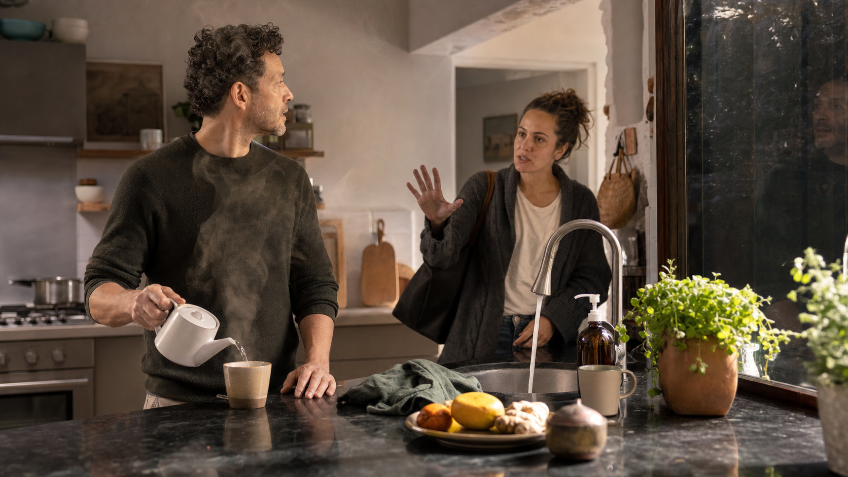 A person preparing tea turns attentively toward a household member who has interrupted them in a sunlit kitchen.