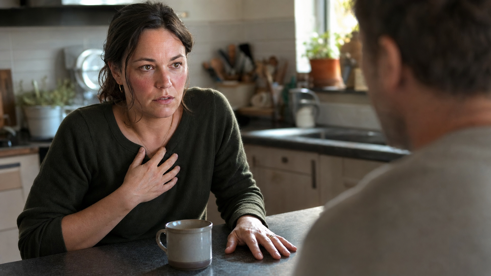 An adult pauses in a kitchen with tense fingers, raised shoulders, and one hand near the chest during a conversation.