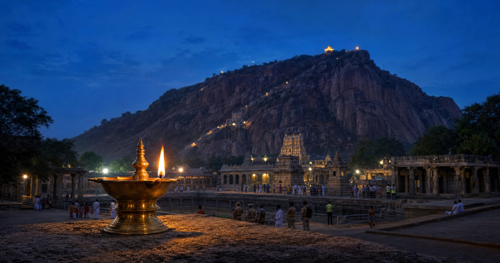 A traditional oil lamp burns at dusk before Thirupparankundram hill as worshippers, temple attendants, safety staff and administrators gather in defined areas.