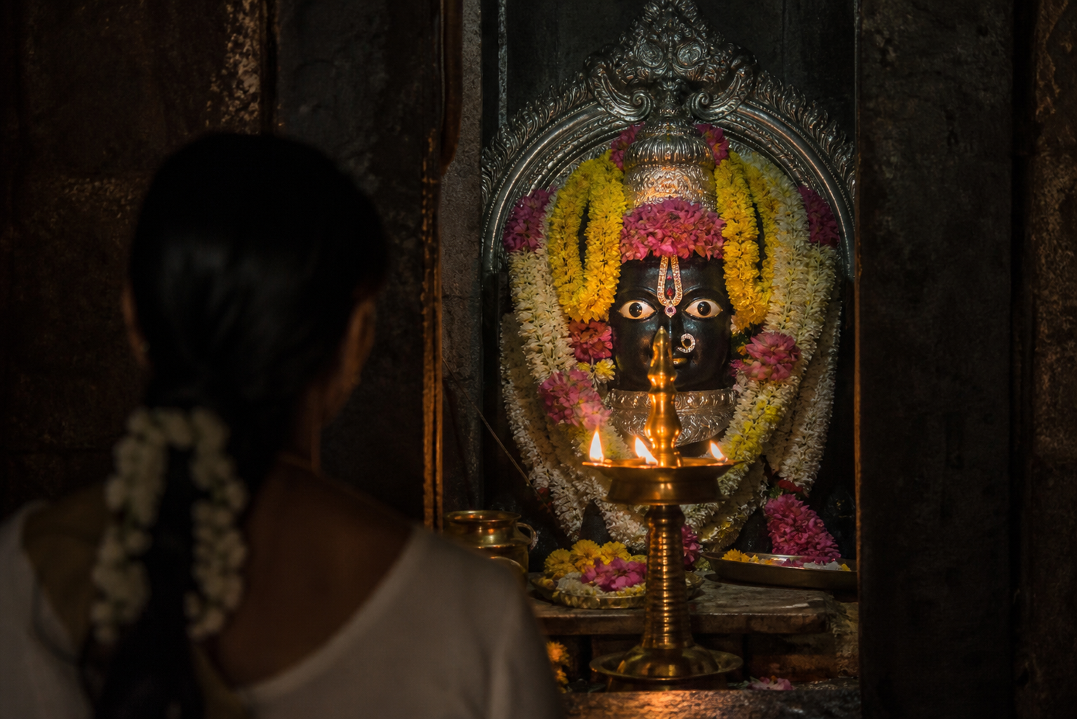 A devotee faces the direct gaze of a deity image across a glowing oil lamp in a Hindu temple sanctum.