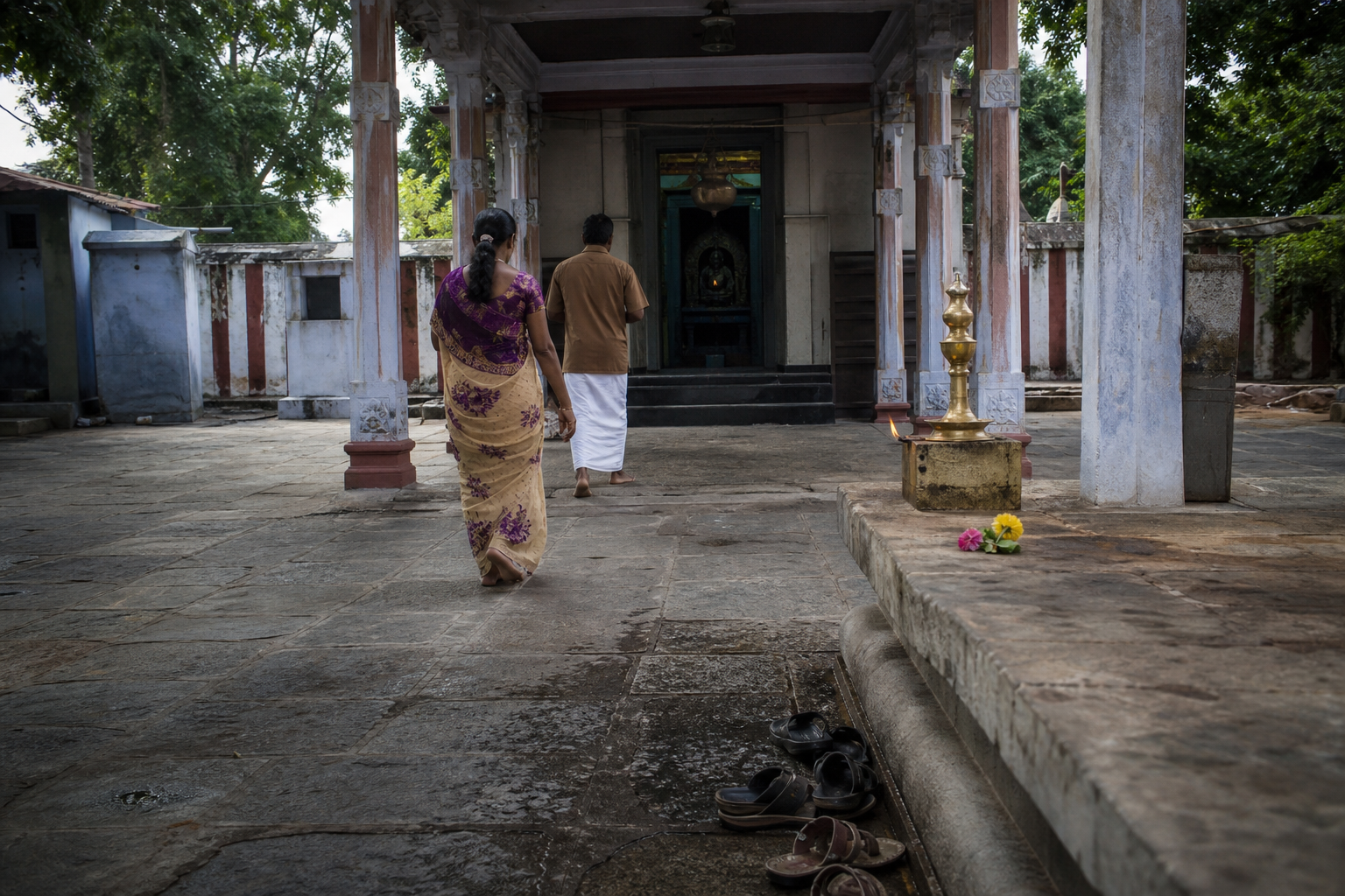 Pilgrims walk through the quiet stone courtyard of a small Tamil Nadu Shiva temple beside a lamp and flower offering.