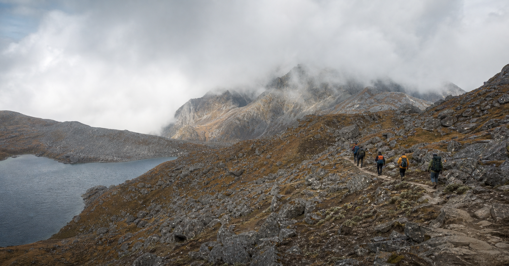 Search-and-rescue personnel walk along a rocky Himalayan trail above a misty alpine lake.