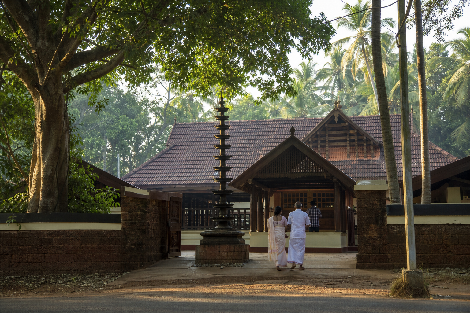 Visitors pause on a tree-shaded path outside a traditional Kerala temple compound with tiled roofs and an oil-lamp pillar.