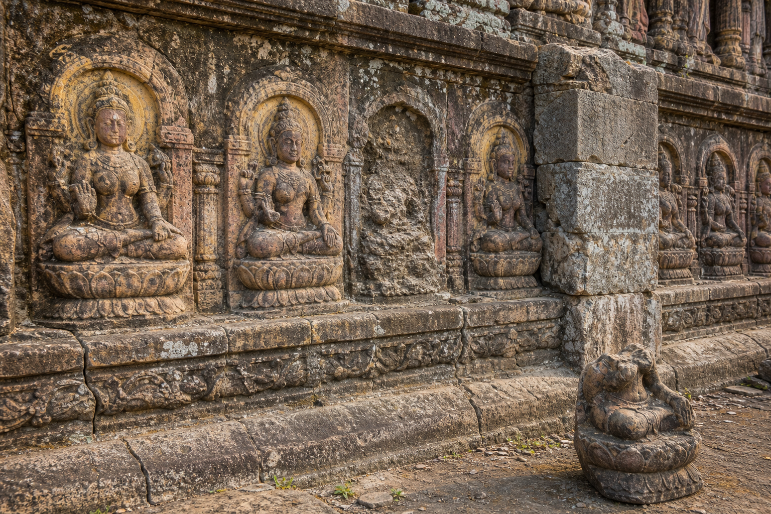 A weathered temple wall contains related goddess niches on a shared plinth, with some carvings eroded or partly obscured.