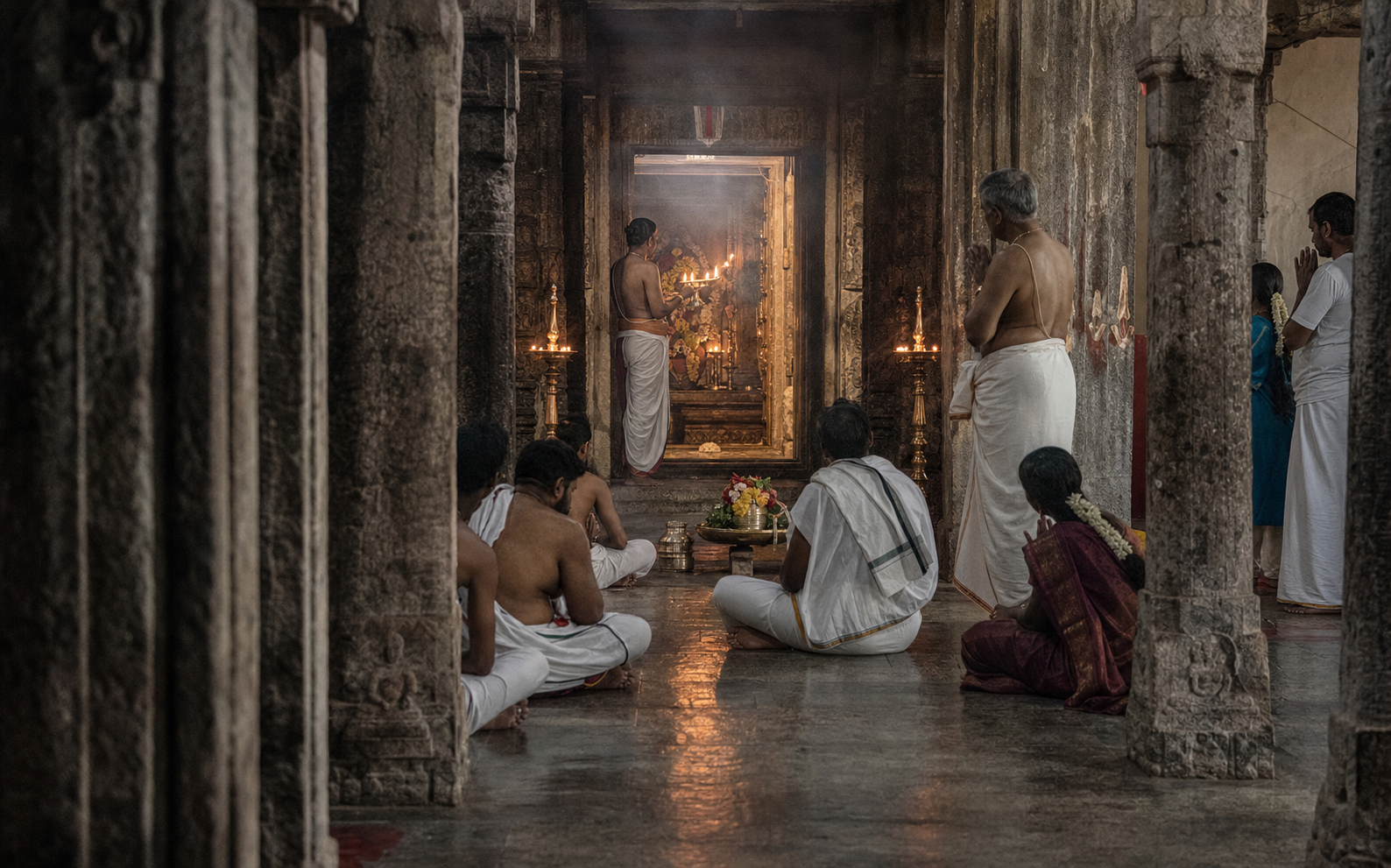 Unshod devotees worship among granite pillars while a priest holds an oil lamp near the temple sanctum.