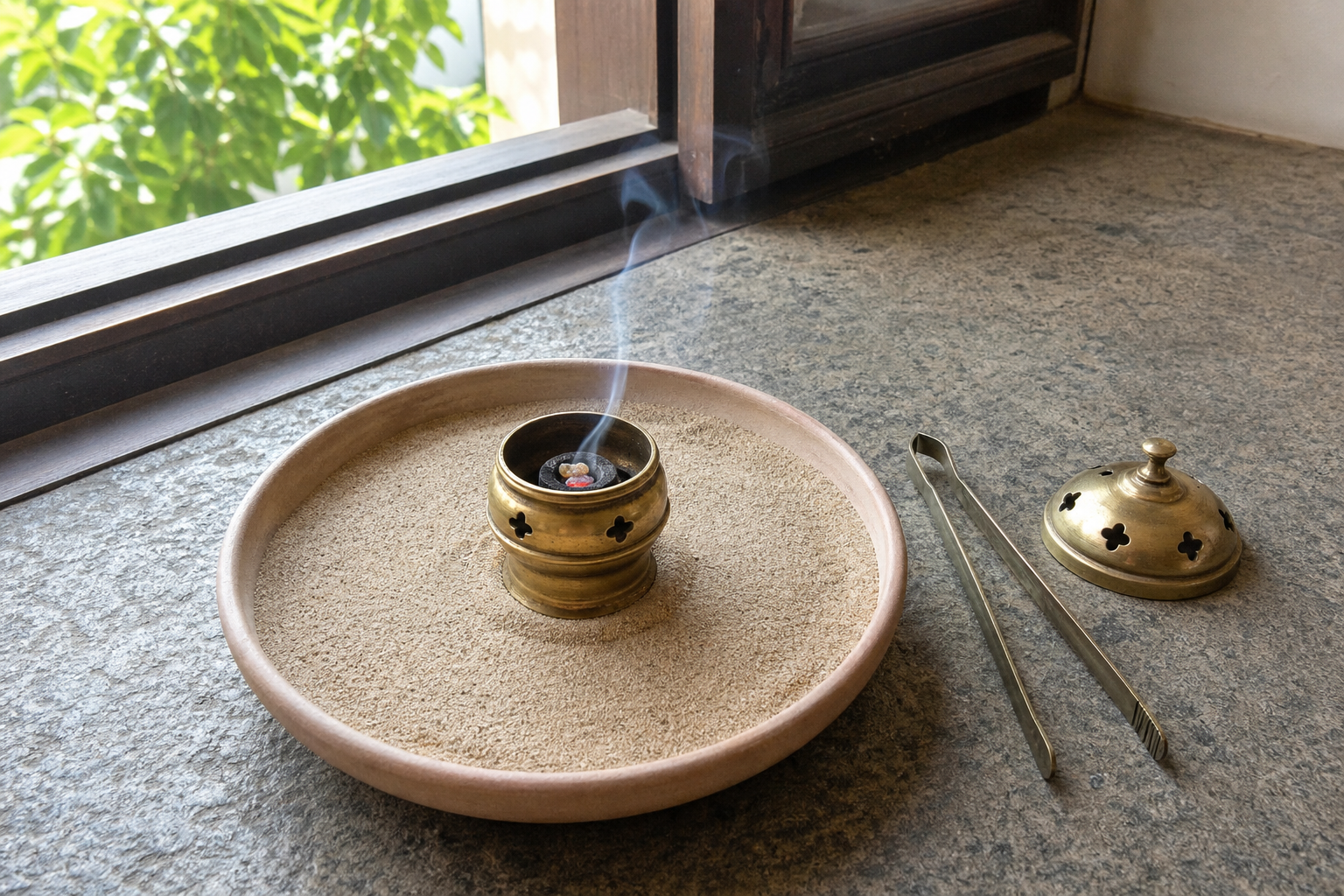 A brass burner rests in a sand-filled ceramic plate near an open window, with tongs and a metal lid beside it and only a thin stream of smoke.