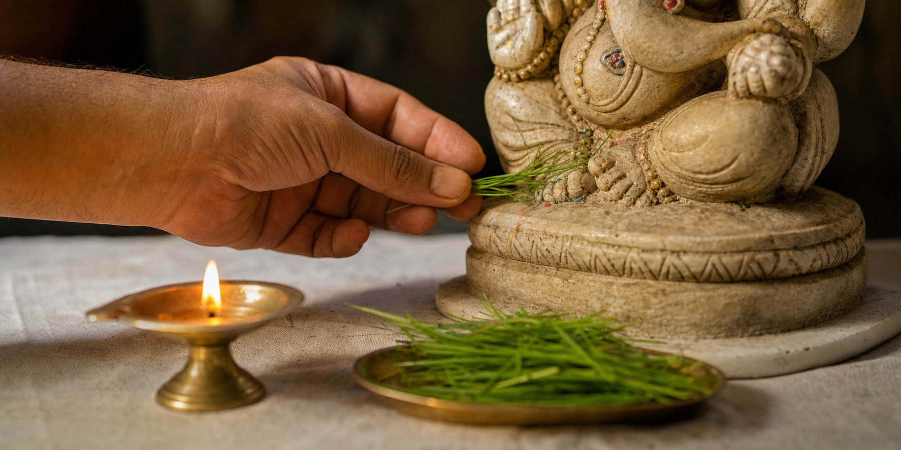 A devotee carefully places a final blade of green durva at the base of a Ganesha murti beside a glowing oil lamp.