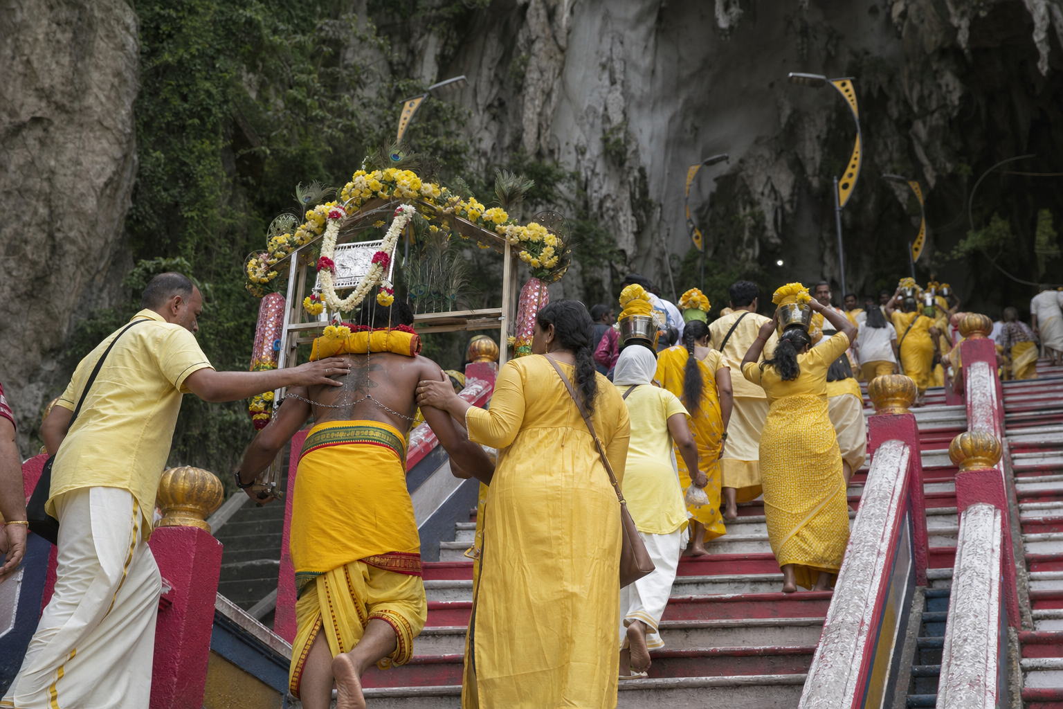 A kavadi bearer supported by family members climbs the Batu Caves staircase among devotees carrying milk pots.