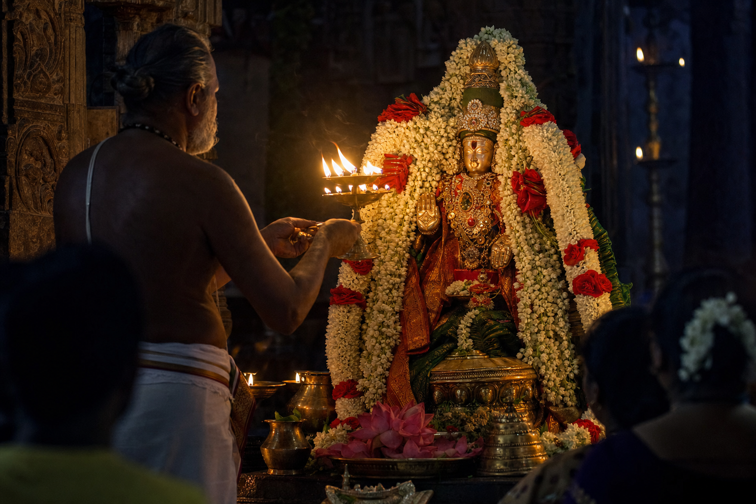 A priest presents a brass flame lamp before a flower-garlanded bronze deity as worshippers stand in the shadowed temple interior.