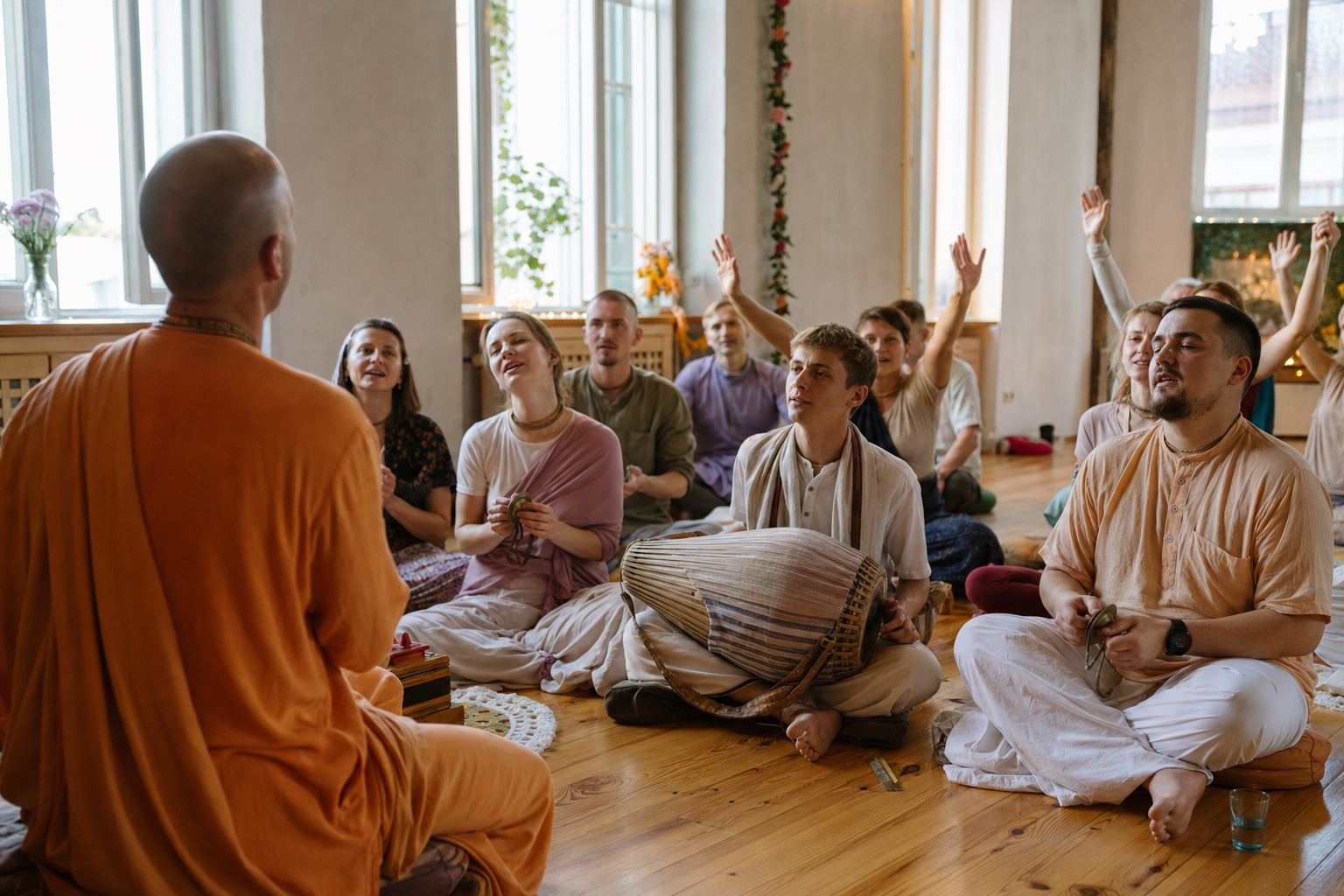 Devotees seated in a Riga hall participate in a Vaishnava kirtan with a drum, hand cymbals, and a saffron-robed leader viewed from behind.