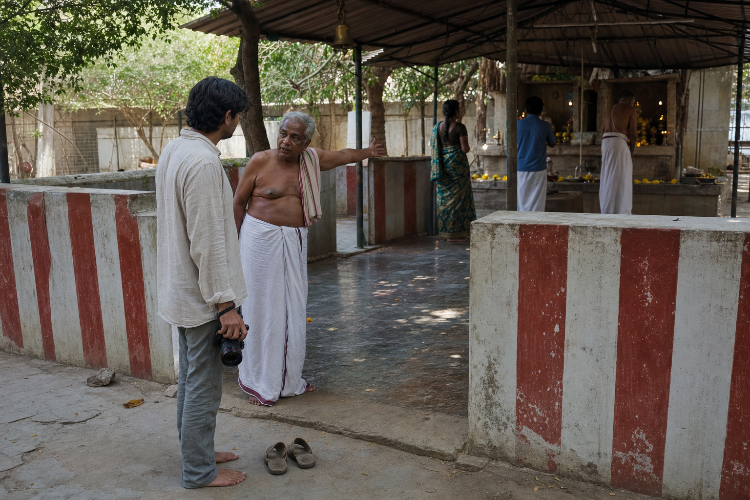 A visitor with a lowered camera speaks with a shrine caretaker beside sandals placed outside the sacred enclosure.