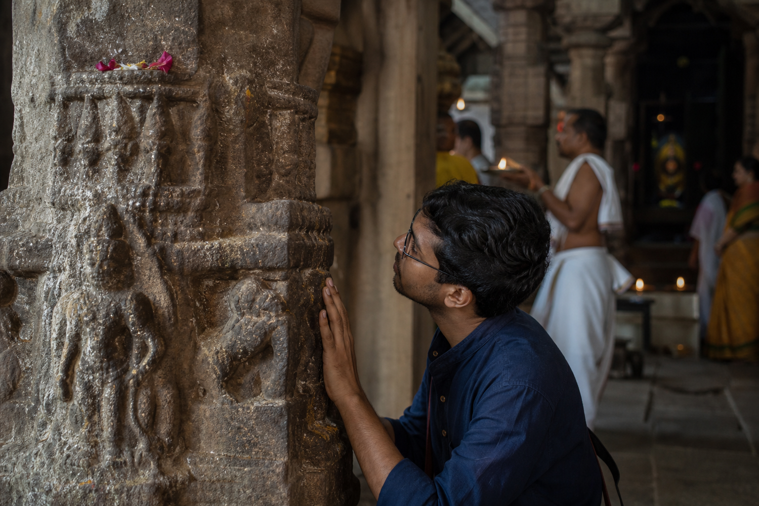 A visitor studies the weathered carvings and tool marks on a stone pillar inside an active Shaivite temple.