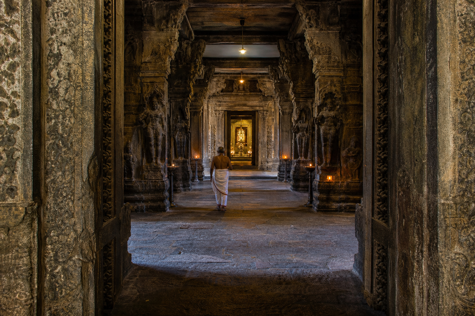 A devotee walks through a pillared mandapam and layered temple thresholds toward a softly lit sanctum doorway.