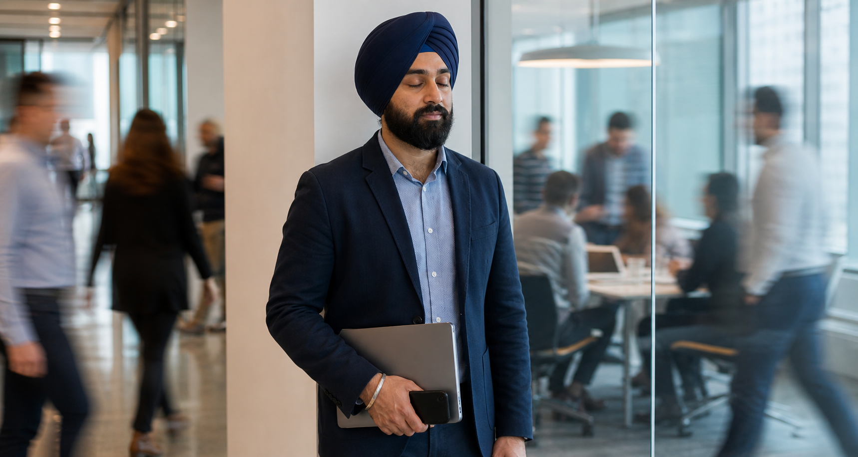 A Sikh professional pauses calmly outside a busy meeting room while coworkers pass in the background.