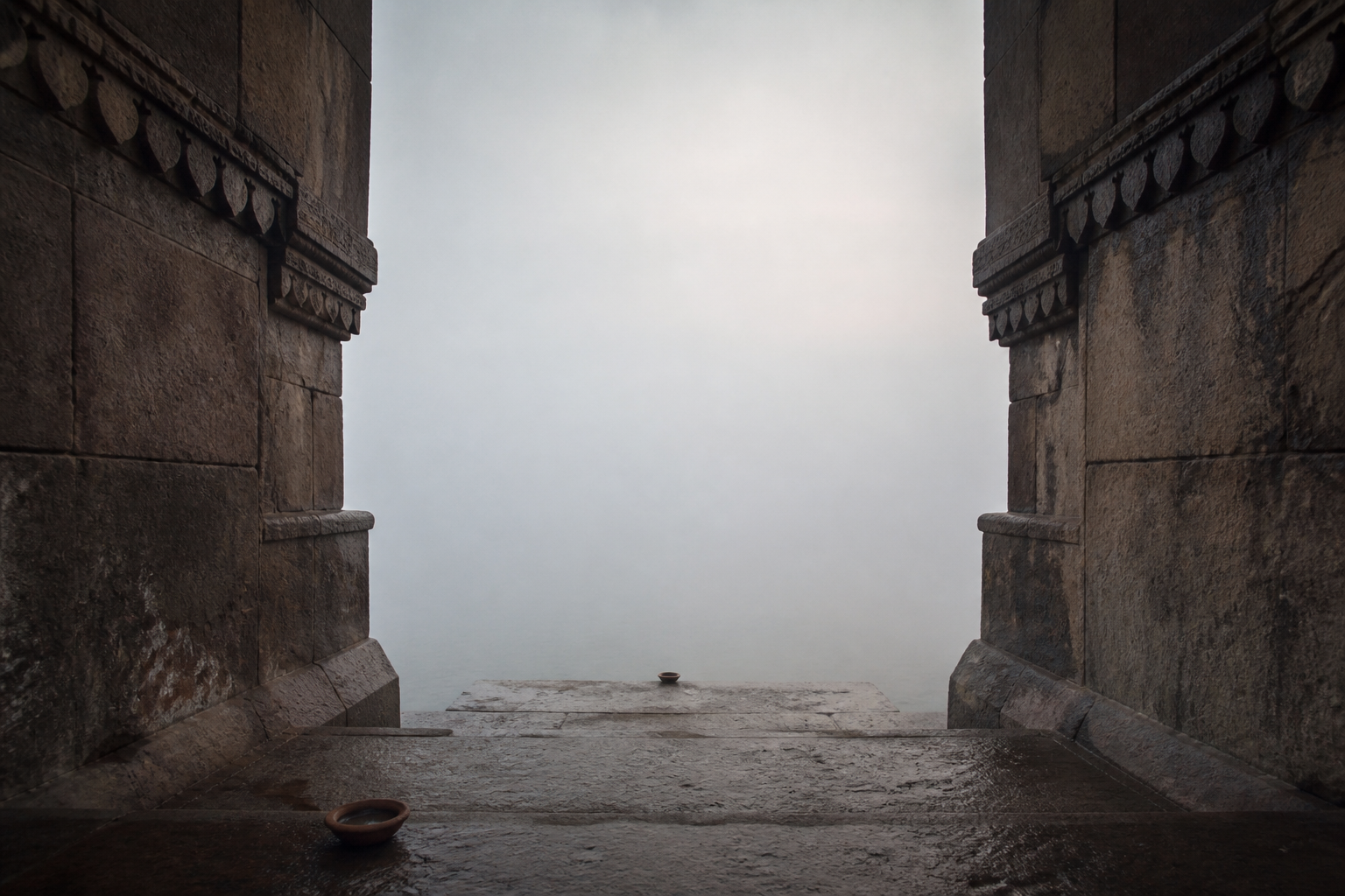 A mist-filled stone passage descends to the Ganga between an extinguished clay lamp and an unlit lamp.