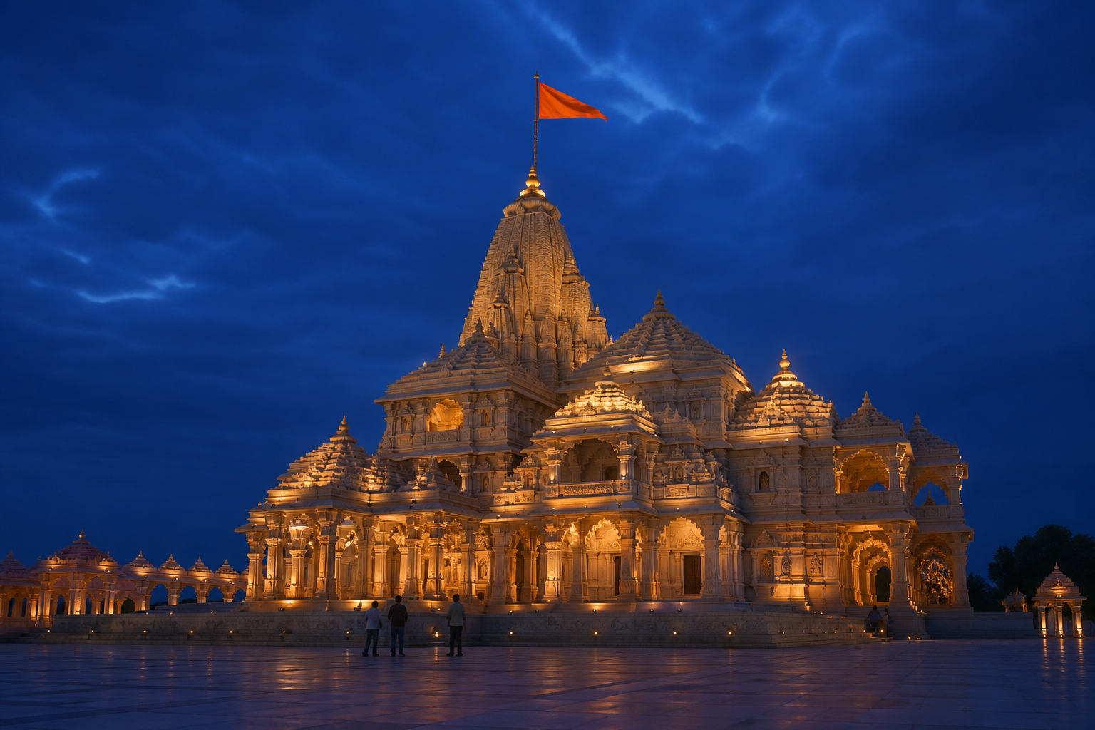 An illuminated carved-stone temple rises above a courtyard at dusk, with a saffron flag flying from its highest central spire and visitors appearing small below.