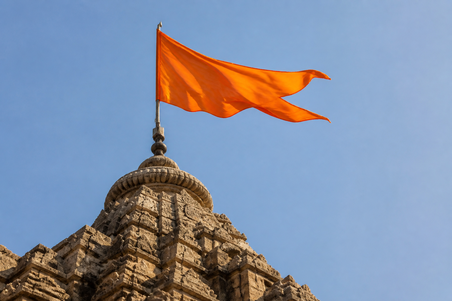 A plain saffron flag billows in morning light above the carved stone spire of a North Indian temple.