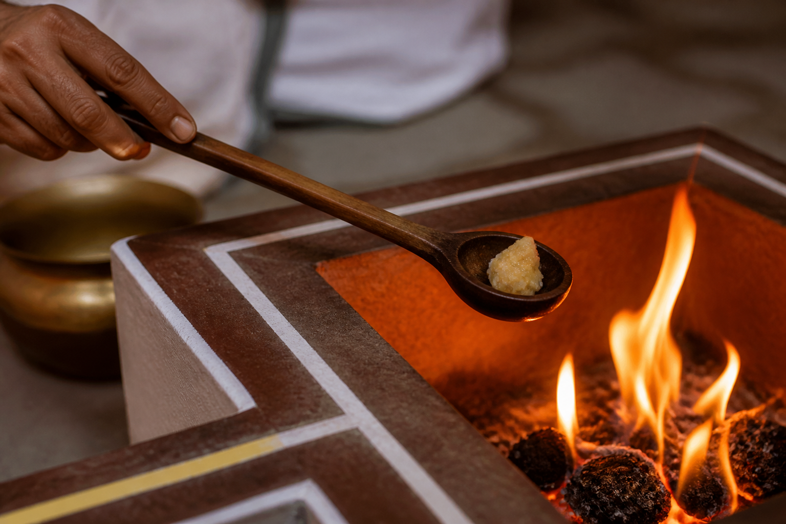 A wooden ladle holds a measured offering over a small ritual fire beside a plain metal vessel.