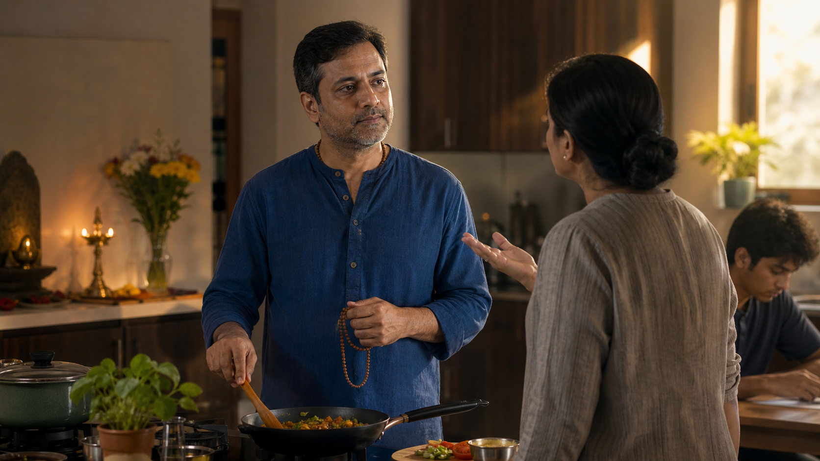 A bhakti practitioner calmly prepares food and listens to a family member beside prayer beads and a small flower-decorated home altar.