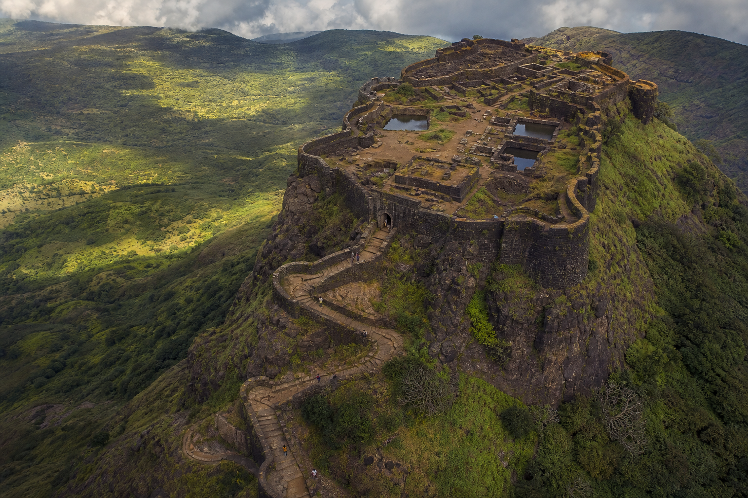 An elevated view shows a hill fort's steep ridge, zigzag approach, recessed gateway, bastions, ramparts, cisterns and interior foundations.