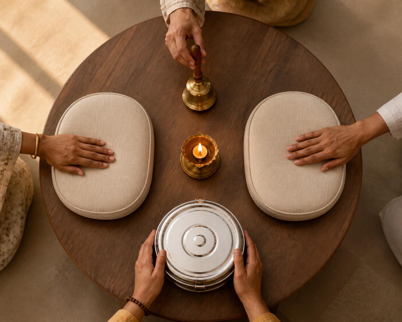 A bell, two cushions, a covered meal container, and a lit oil lamp form a circle on a wooden table as several people tend the objects together.