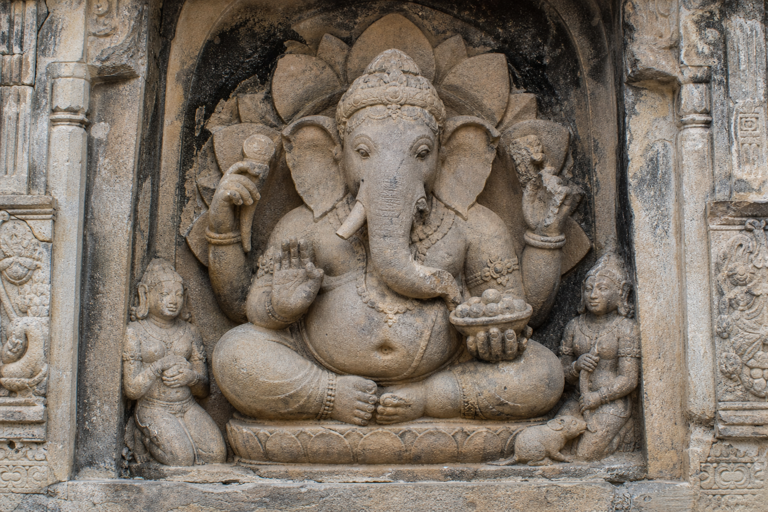 Weathered stone relief of seated Ganesha with multiple arms, attendants, lotus ornament, and a small mouse at the base of the niche.