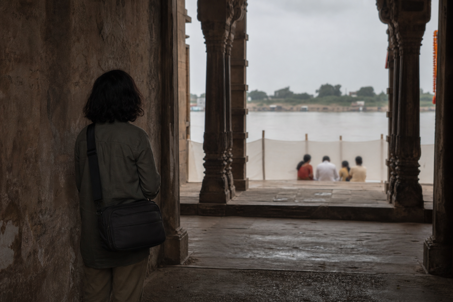 A visitor waits at a distance with a camera stored away while a screened family gathering takes place beside a river.