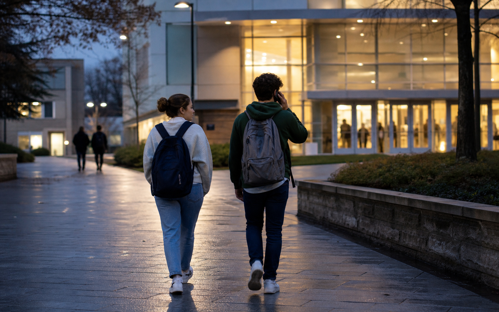 A student and supportive friend move toward a brightly lit campus lobby while the student makes a phone call.