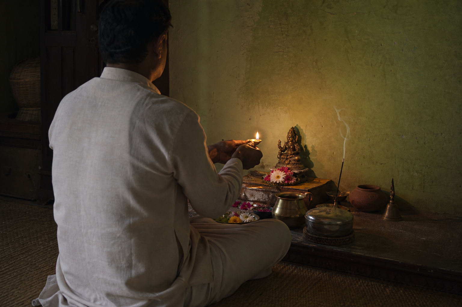 A seated devotee holds a small oil lamp before a simple home altar with the other puja offerings nearby.
