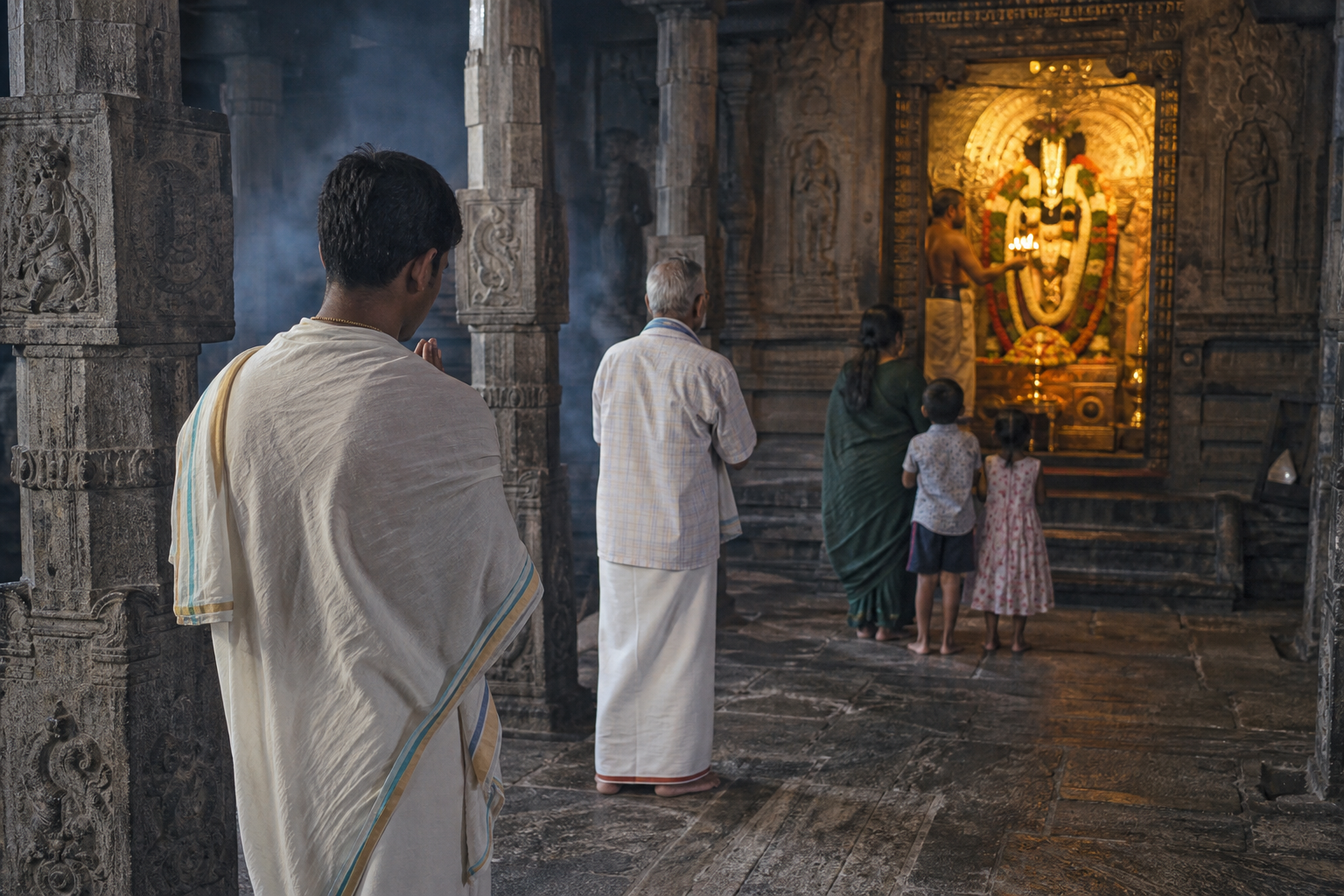 A pilgrim waits with joined hands at a temple sanctum while a priest offers a lamp before an adorned Vishnu murti.
