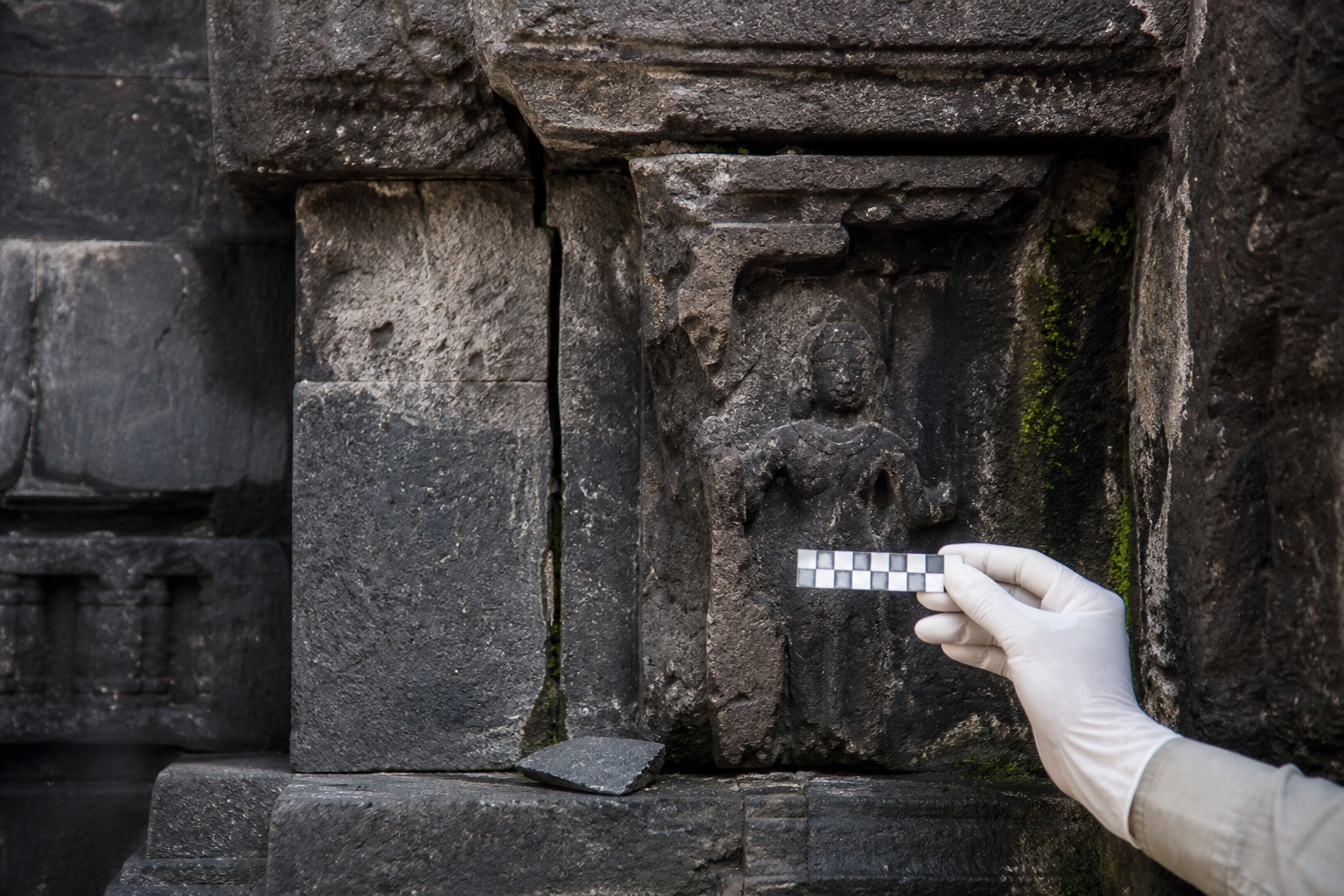 Close view of a cracked and moisture-stained carved stone wall while a gloved conservator documents the damage without touching it.