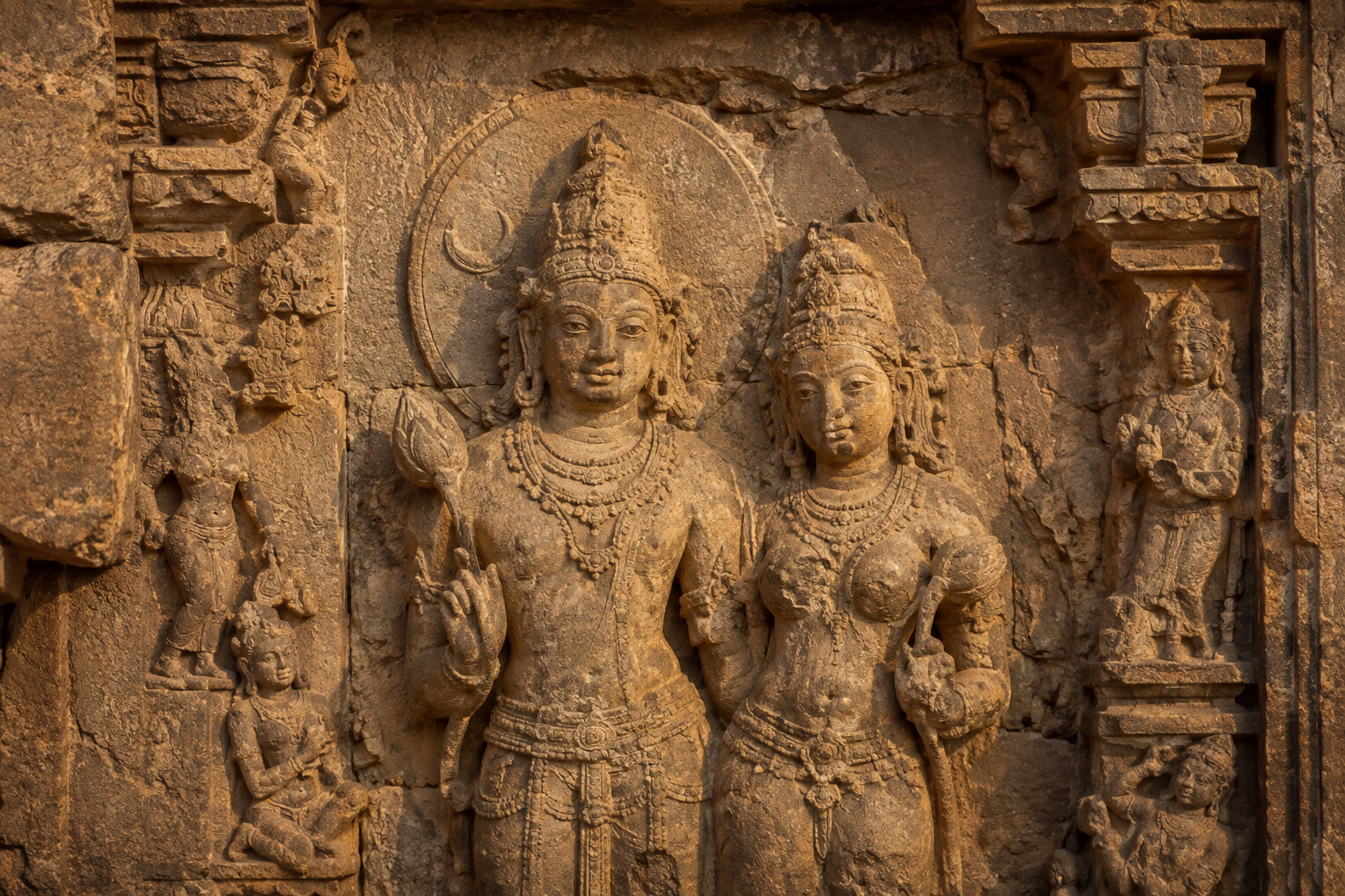 Weathered sandstone temple relief showing a haloed male celestial deity beside a female companion, with fragmentary lotus stems and smaller celestial figures around them.