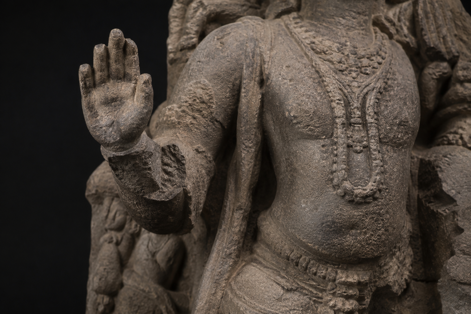 Close view of a weathered stone figure with a raised hand, damaged forearm, and incomplete surrounding carvings.