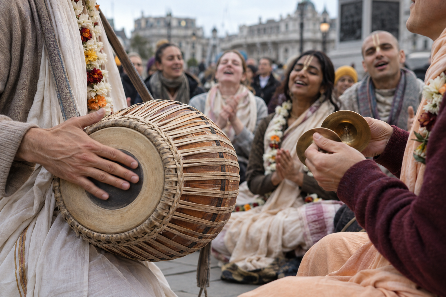 A devotee plays a double-headed drum beside hand cymbals while a circle of singers participates in outdoor kirtan.