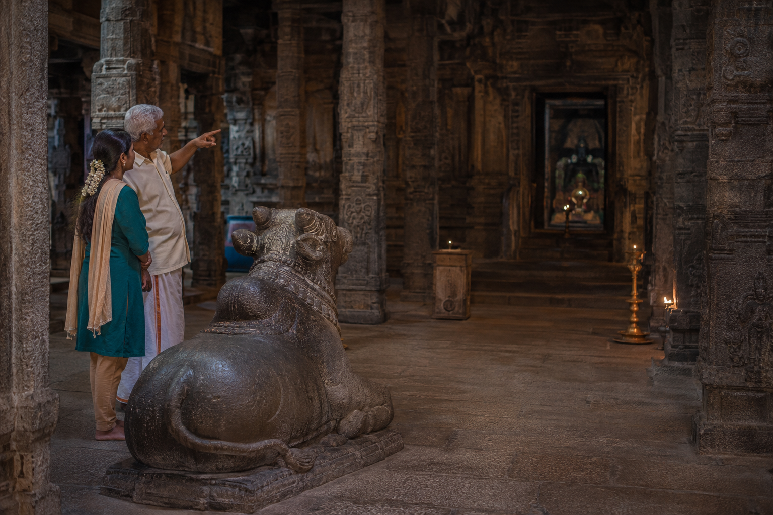 An older pilgrim gestures toward the off-axis stone Nandi while a younger companion listens inside a lamp-lit temple hall.