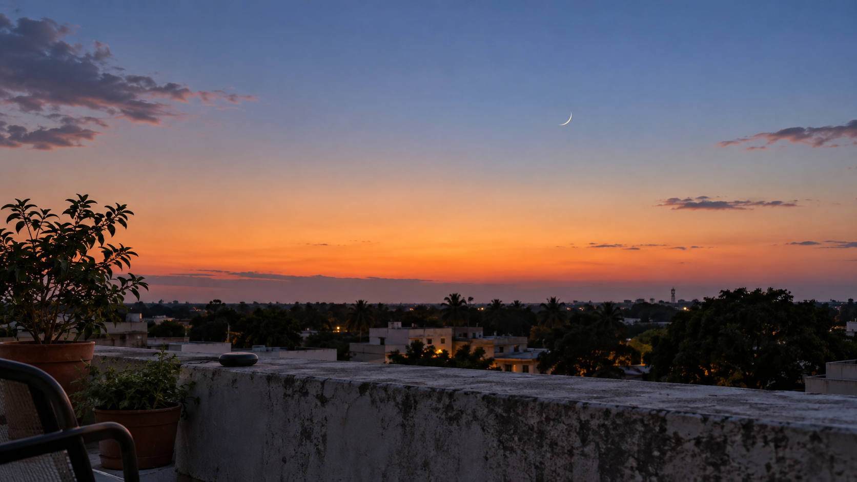 A thin crescent moon appears above a clear western horizon viewed from a residential rooftop at dusk.